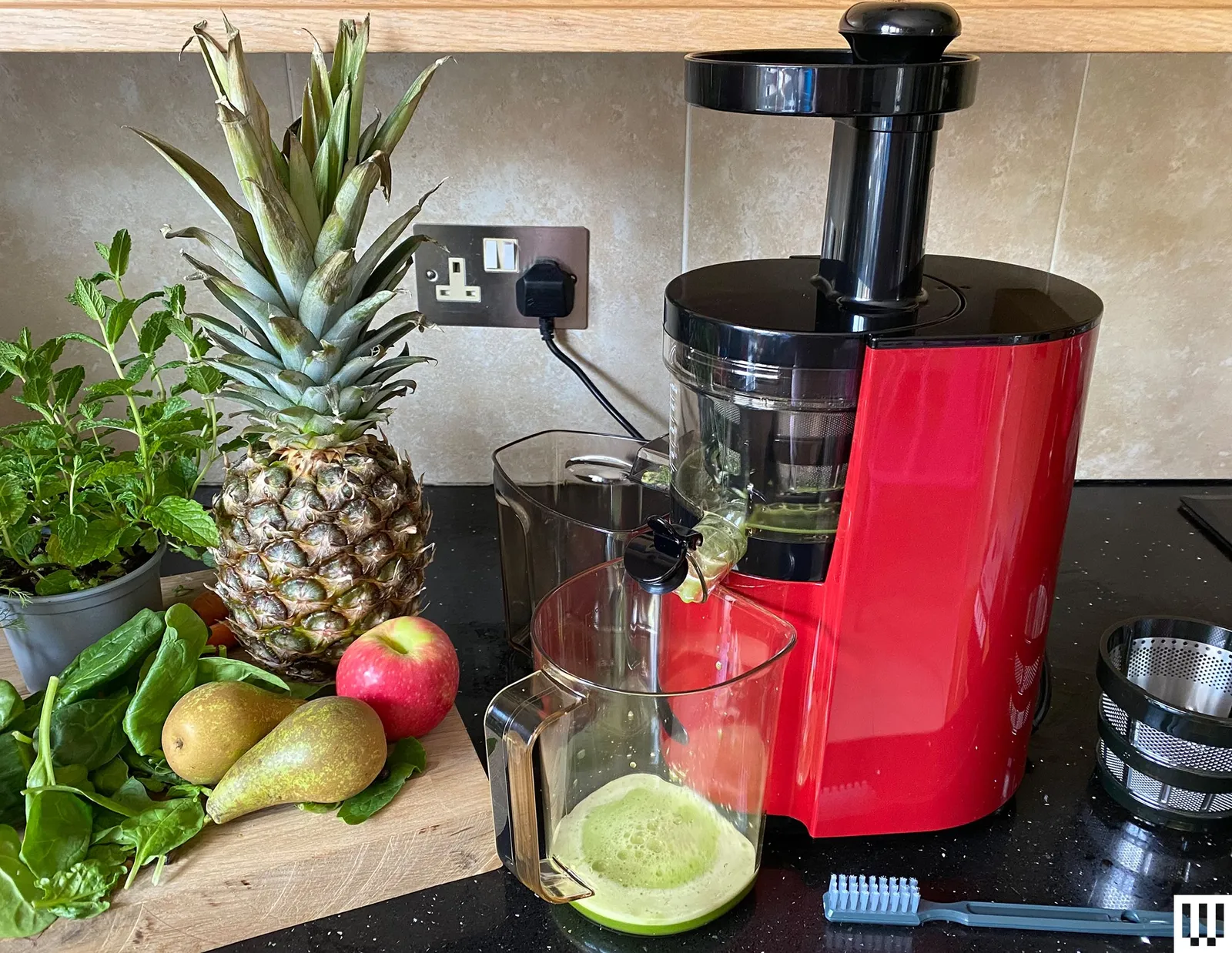 Kitchen counter that has a cutting board on the left with fruits and vegetables and a red cylindrical juicing machine on...