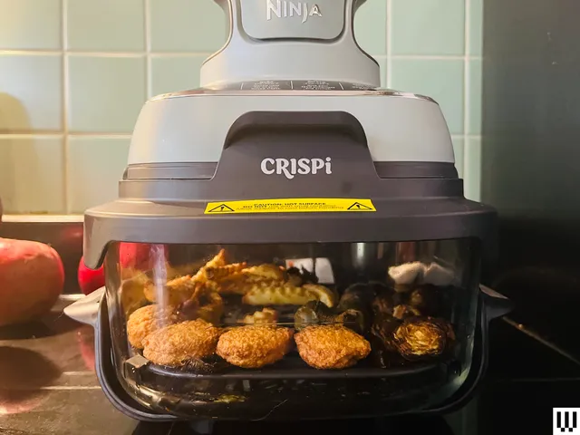 Closeup of the Ninja Crispi, a portable airfryer comprised of a clear dish on the bottom and a two-tier funnel-shaped device attached on top, sitting on a kitchen counter with chicken nuggets and french fries inside the container