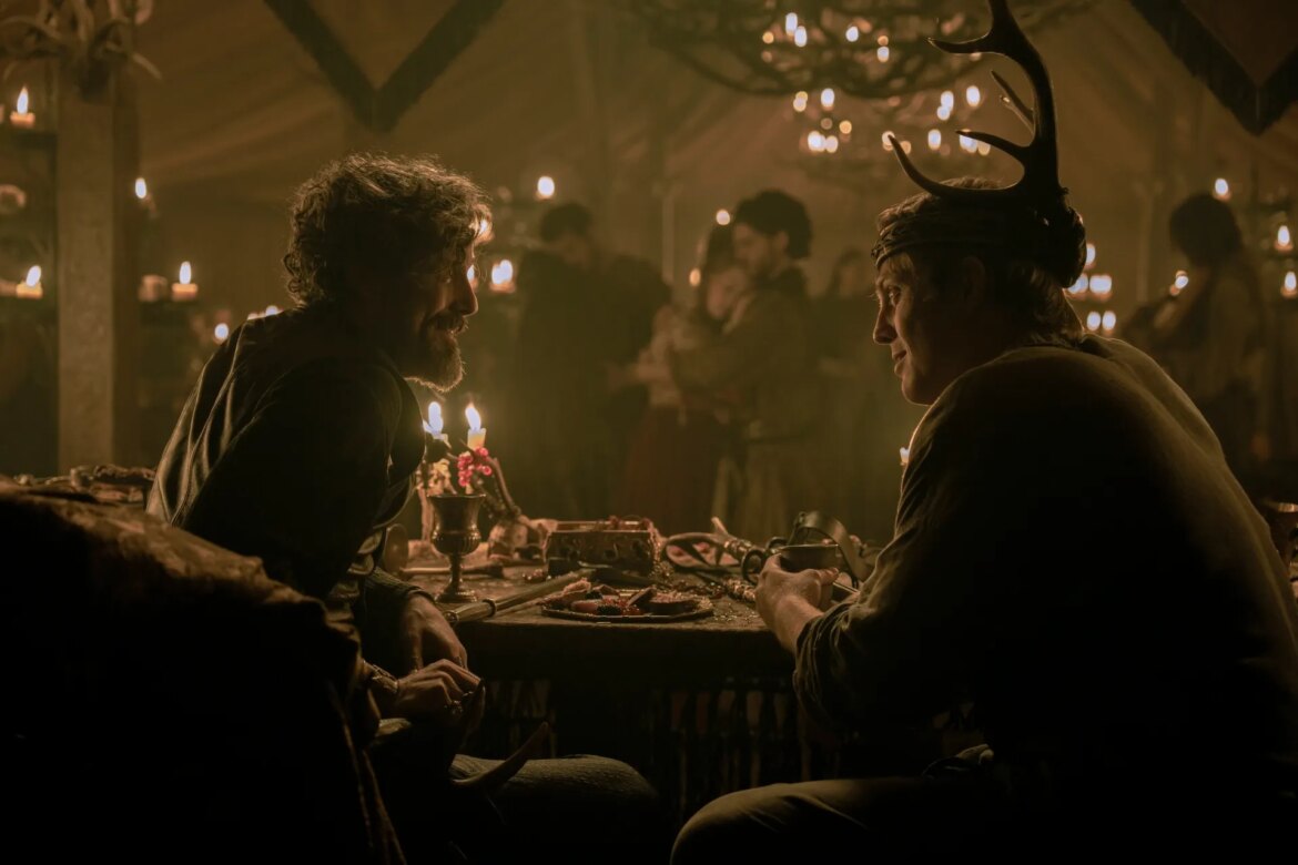 Two men sitting together at a table in room where a party is being held. The man on the right is wearing a crown adored with antlers.