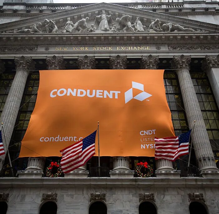 Conduent Inc. signage is displayed as American flags fly outside of the New York Stock Exchange (NYSE) in New York, U.S., on Tuesday, Jan. 3, 2017. U.S. stocks rose Tuesday, pointing to the biggest gain in a month for Wall Street