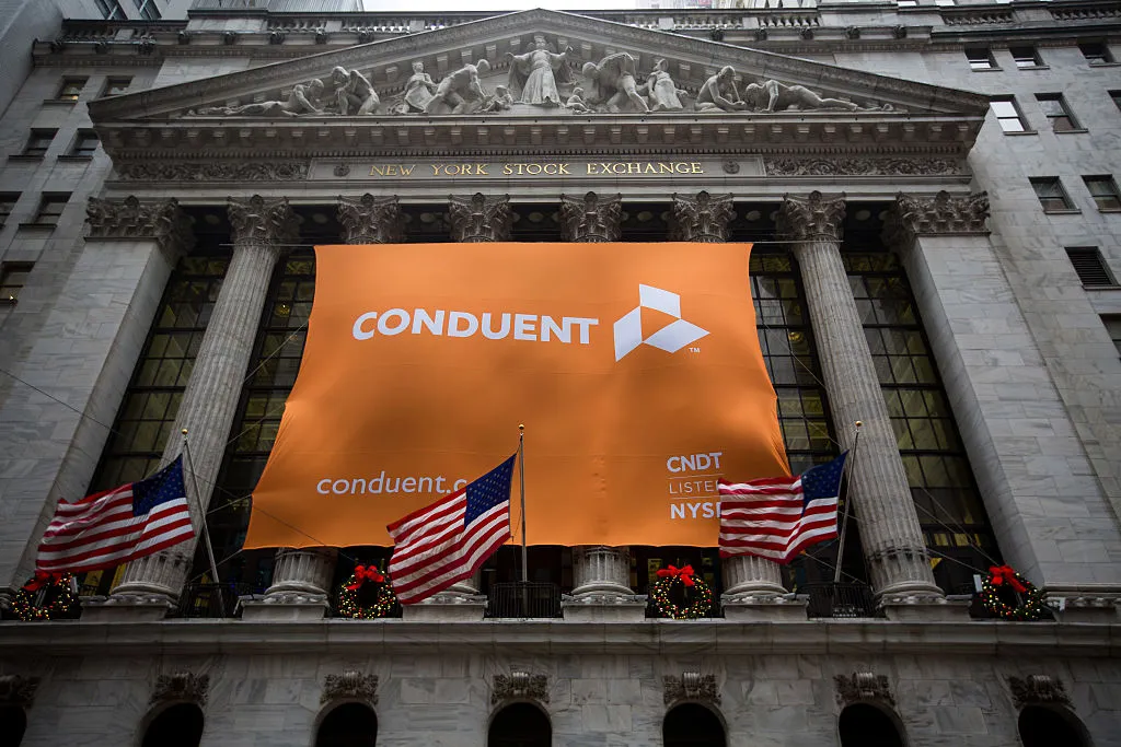 Conduent Inc. signage is displayed as American flags fly outside of the New York Stock Exchange (NYSE) in New York, U.S., on Tuesday, Jan. 3, 2017. U.S. stocks rose Tuesday, pointing to the biggest gain in a month for Wall Street