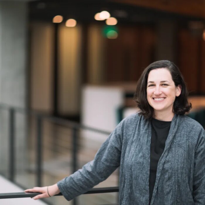 A dark-haired woman poses by a balcony in a gray cardigan sweater.