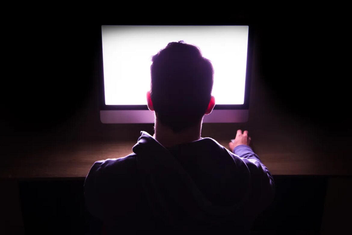 Back view of a man in front of bright computer working in the dark.