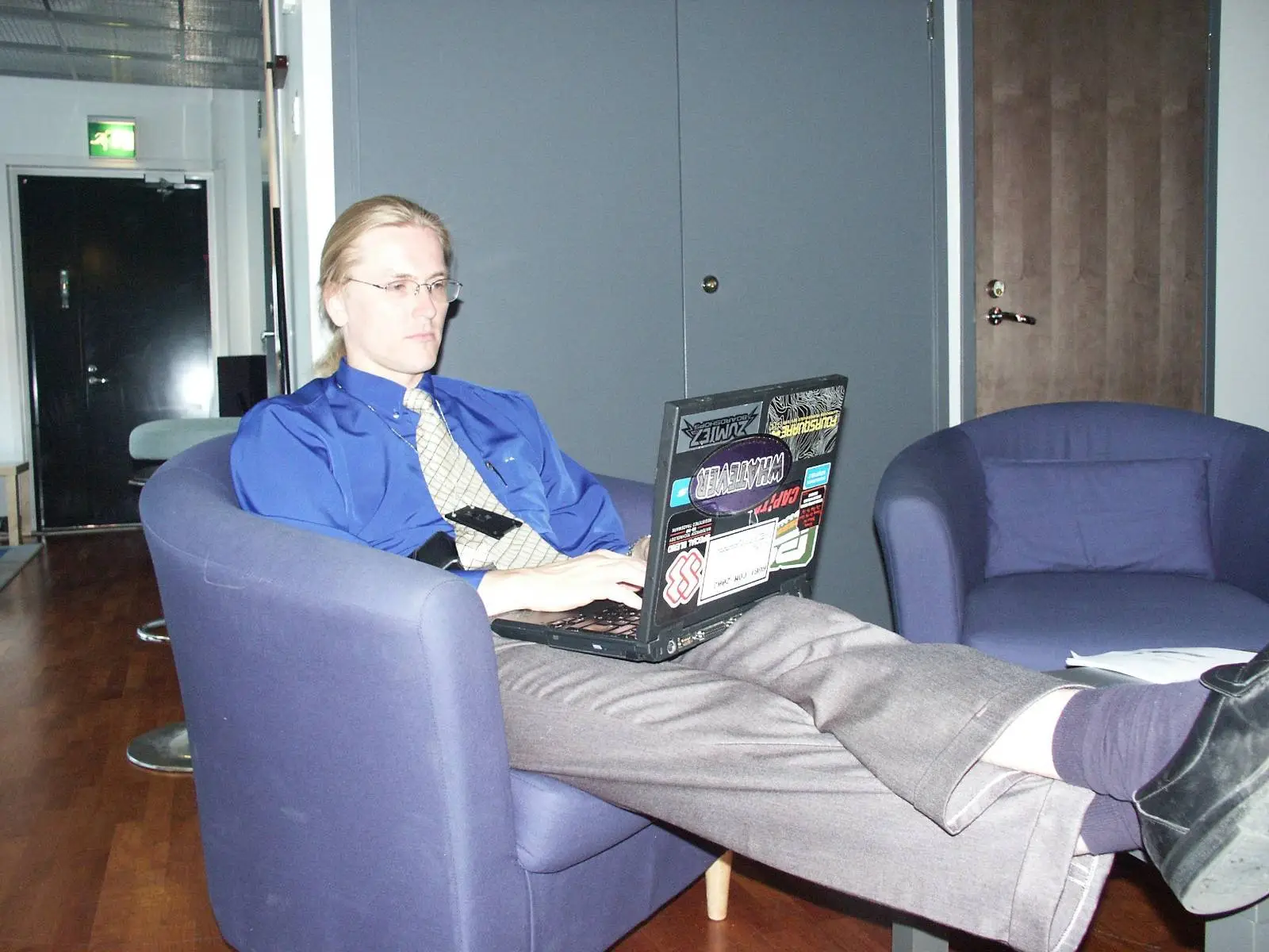 a photo of a younger Mikko Hyppönen, wearing a blue shirt and tie, on a purple chair, with his feet up and a laptop with stickers on his lap.