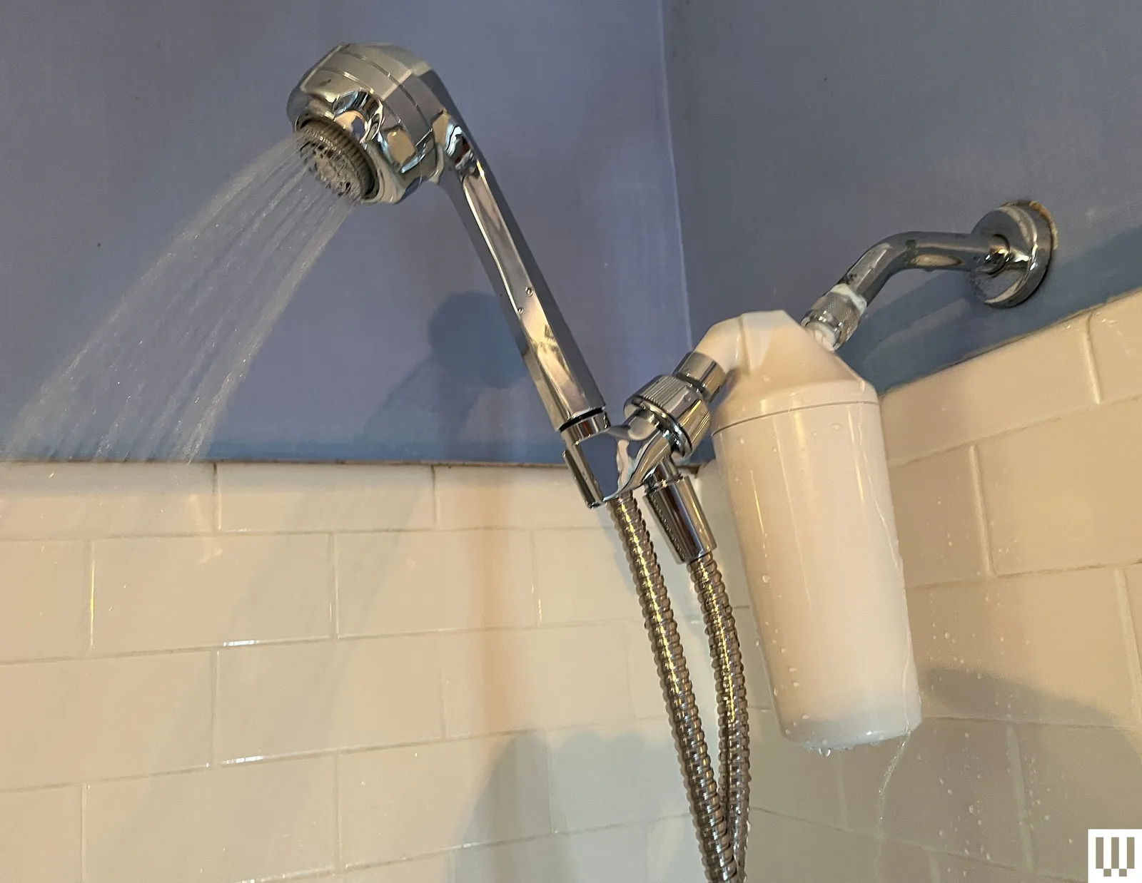Silver elongated showerhead turned on with white tiles in the background and a white filter attachment connected to the...