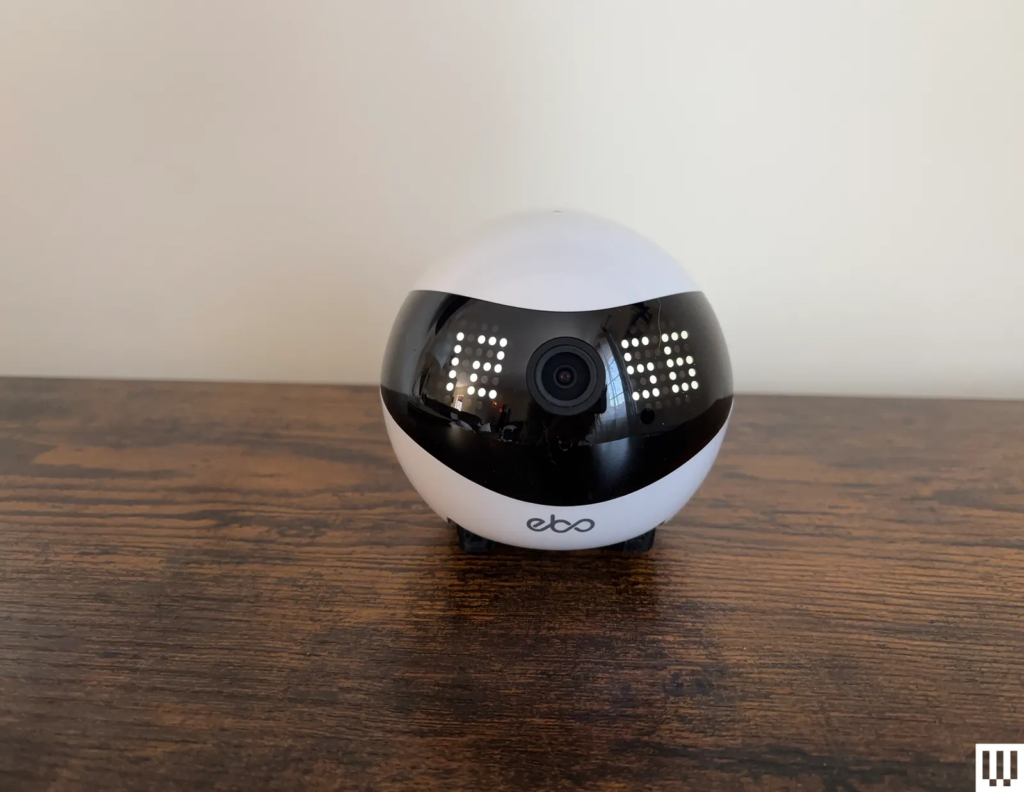 White pet camera on wheels with clock displaying 1256 on face sits on wood ledge with cream wall background