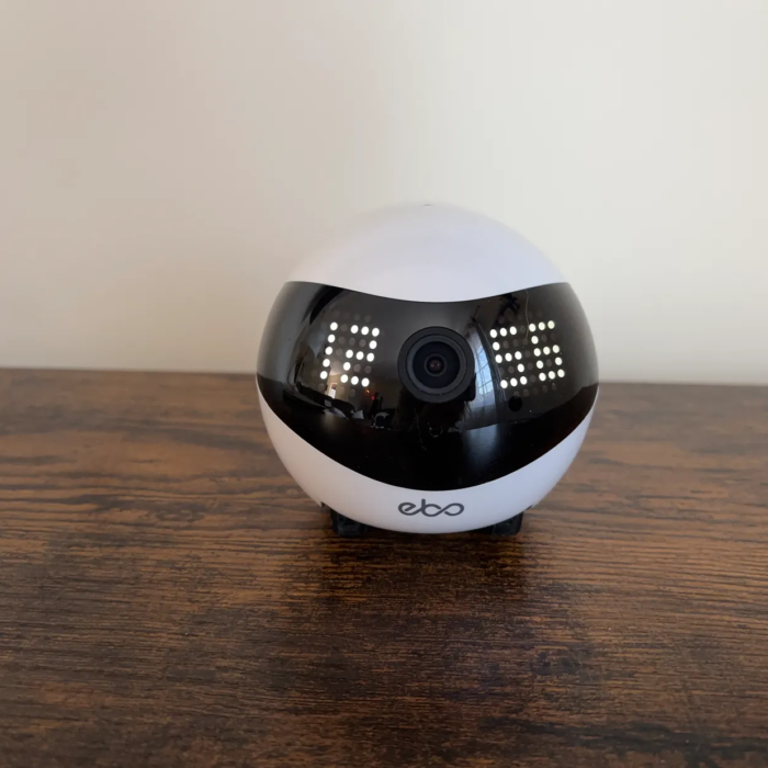 White pet camera on wheels with clock displaying 1256 on face sits on wood ledge with cream wall background