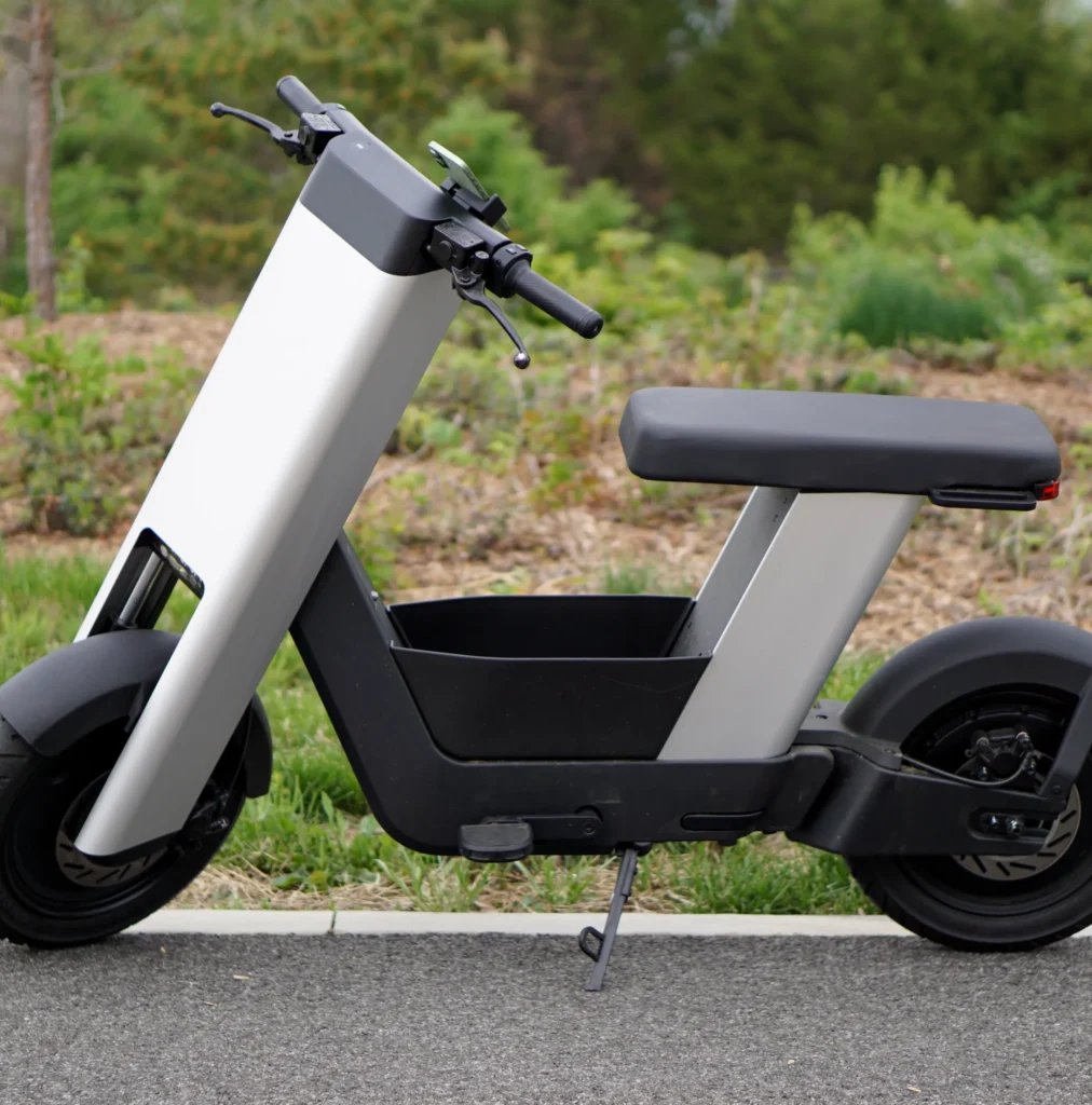 A photo of a silver e-bike from the side, in front of trees.