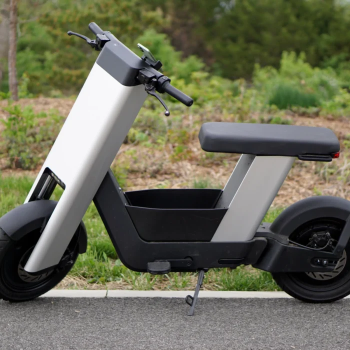 A photo of a silver e-bike from the side, in front of trees.