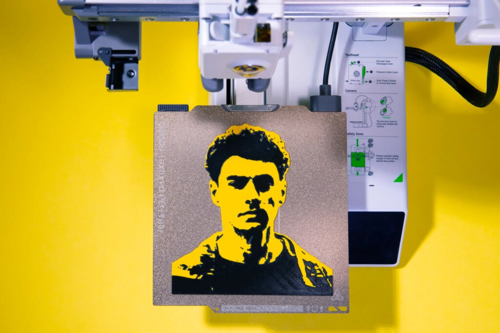 Black and yellow 3D print of Luigi Mangione sitting on a 3D print bed.