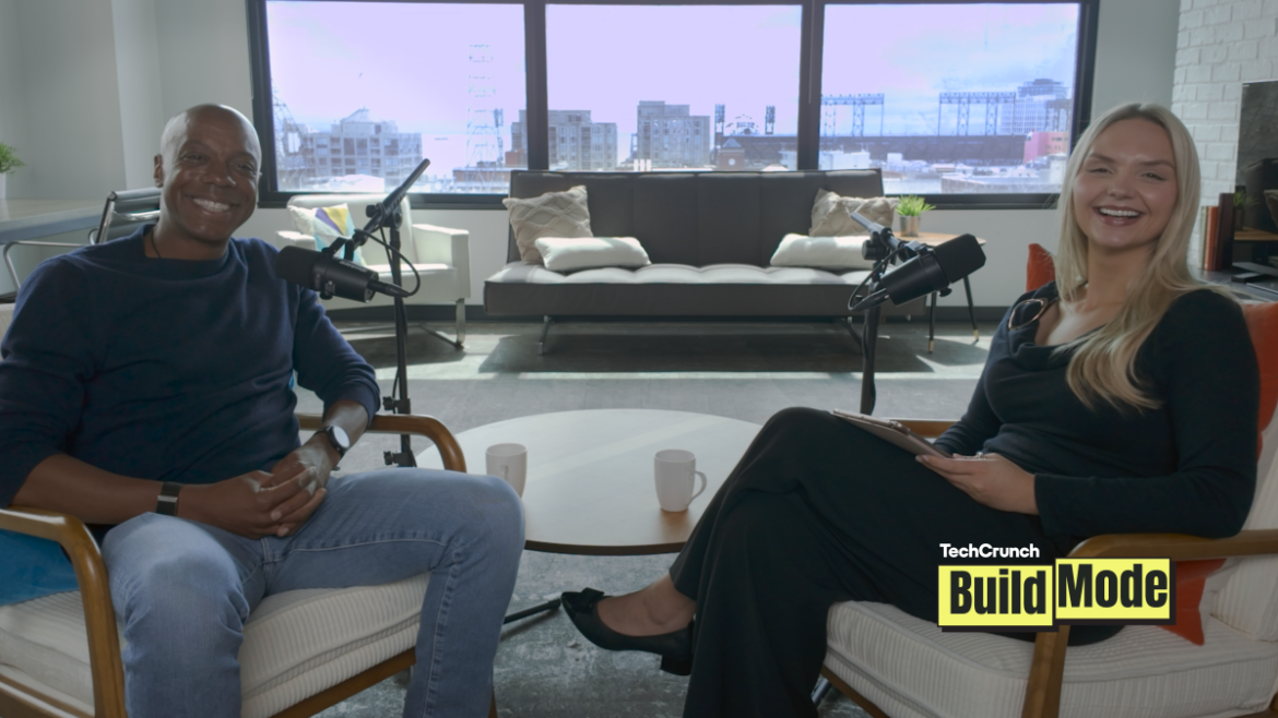 TechCrunch Build Mode podcast host Isabelle Johannessen sits across from venture capitalist Charles Hudson in a bright podcast studio. Both are smiling toward the camera, seated in armchairs with microphones positioned between them. A coffee table with two mugs sits in the foreground, while large windows behind them overlook the San Francisco skyline. The TechCrunch Build Mode logo appears in the bottom-right corner.