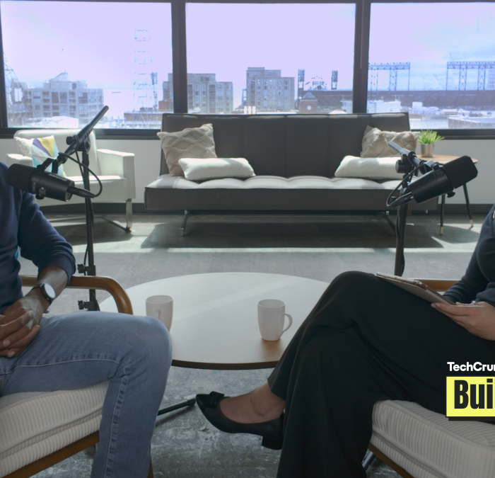 TechCrunch Build Mode podcast host Isabelle Johannessen sits across from venture capitalist Charles Hudson in a bright podcast studio. Both are smiling toward the camera, seated in armchairs with microphones positioned between them. A coffee table with two mugs sits in the foreground, while large windows behind them overlook the San Francisco skyline. The TechCrunch Build Mode logo appears in the bottom-right corner.
