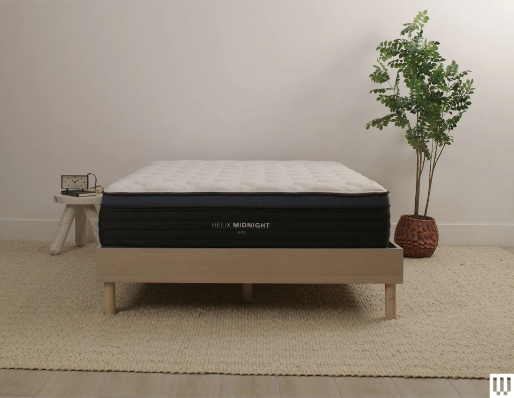Helix Sleep Midnight Luxe a white mattress with blue trim on a minimalist wooden frame with a nightstand and potted...