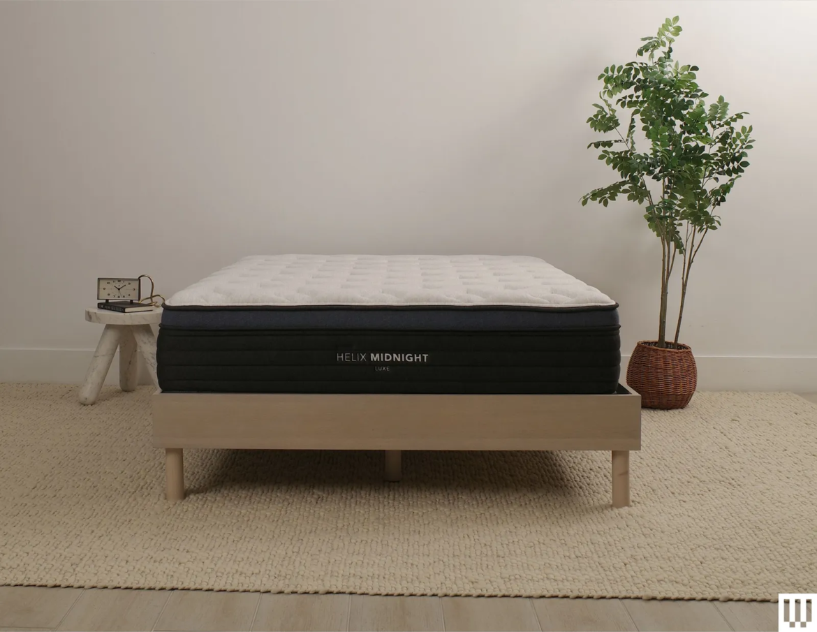Helix Sleep Midnight Luxe a white mattress with blue trim on a minimalist wooden frame with a nightstand and potted...
