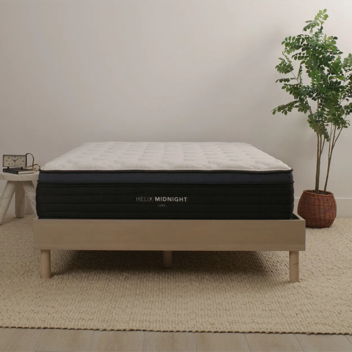 Helix Sleep Midnight Luxe a white mattress with blue trim on a minimalist wooden frame with a nightstand and potted...